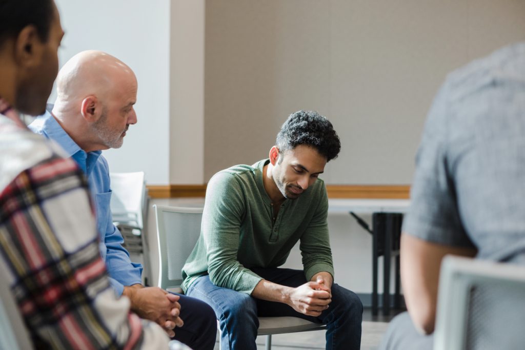 During group therapy, the young adult man looks down at the floor as he takes time to gather his thoughts.